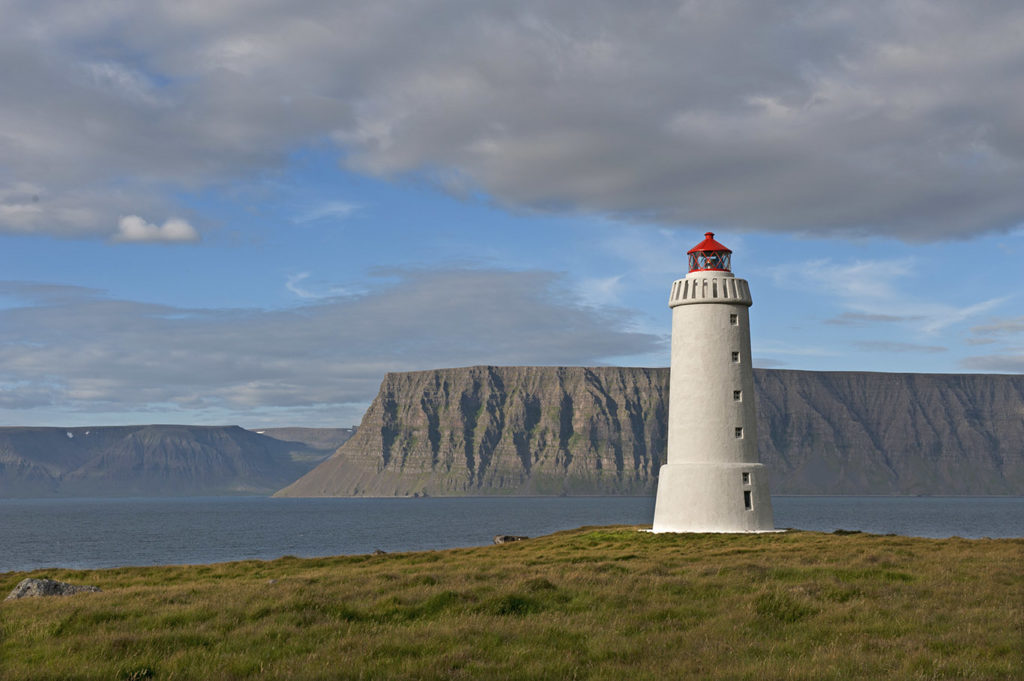 Faro bianco e rosso di Skálavíkurviti con vista sulla baia e montagne sullo sfondo, vicino a Bolungarvík, Islanda.