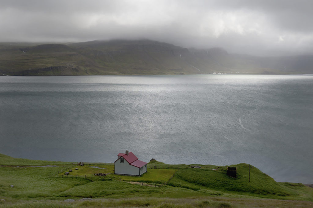 Casetta rossa isolata sulle rive del fiordo Arnarfjörður con cielo grigio e montagne sullo sfondo, Westfjords Islanda.