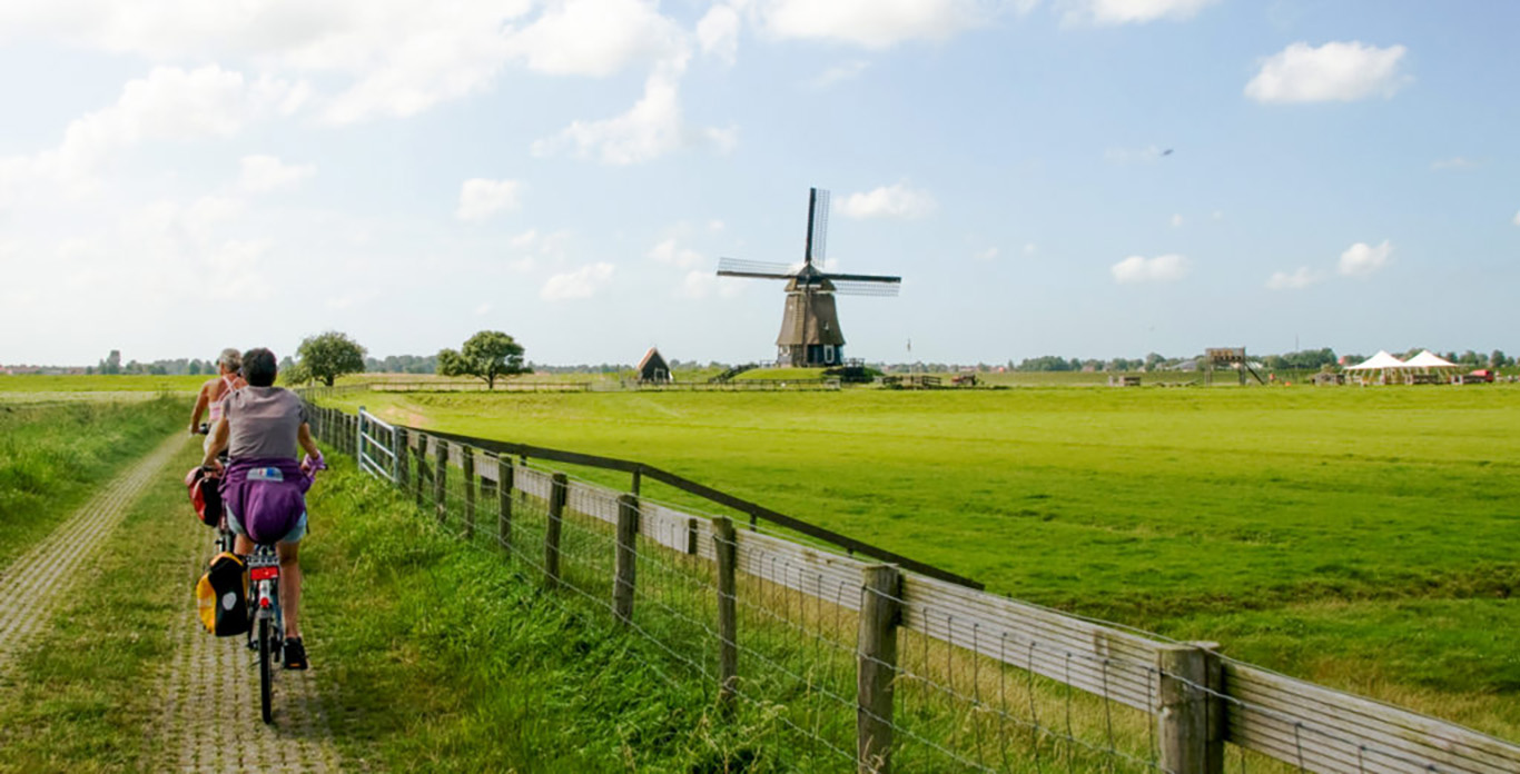 Two cyclists ride on a bicycle path in Holland through a green landscape with a windmill.