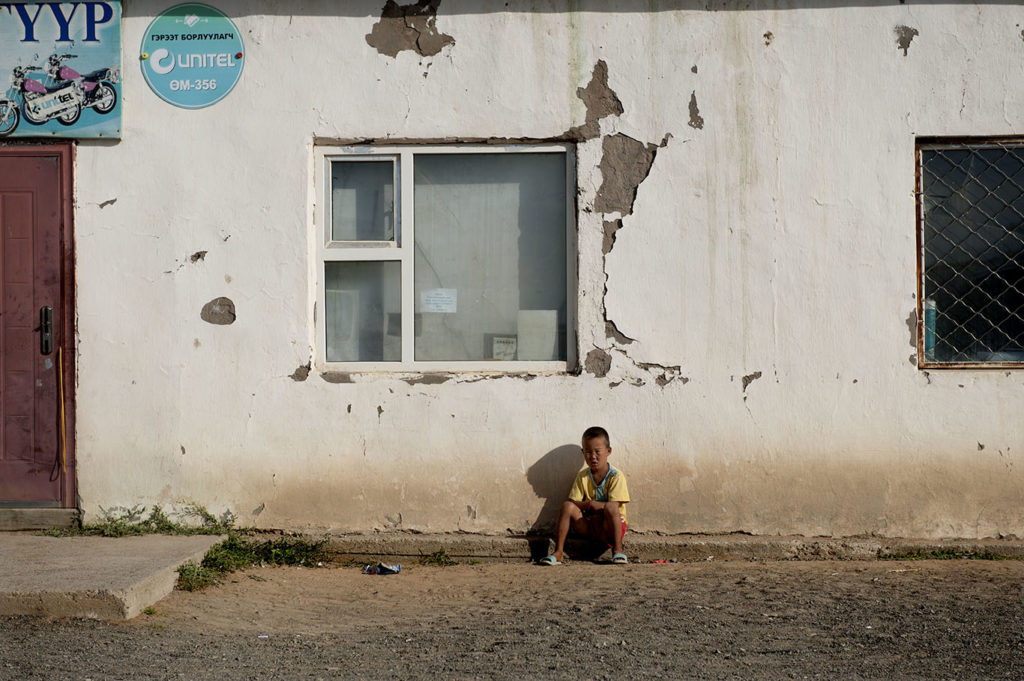 Bambino mongolo seduto accanto a un muro scrostato di un edificio in un villaggio rurale
