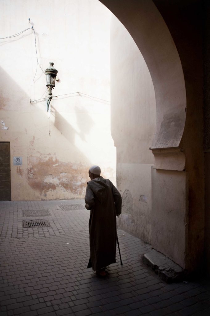 Anziana che cammina in un vicolo illuminato di Marrakech con pareti color sabbia e lanterna appesa.