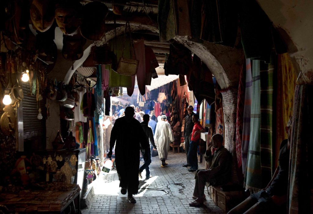 Corridoio ombreggiato nel souk della medina di Marrakech con sagome di passanti.