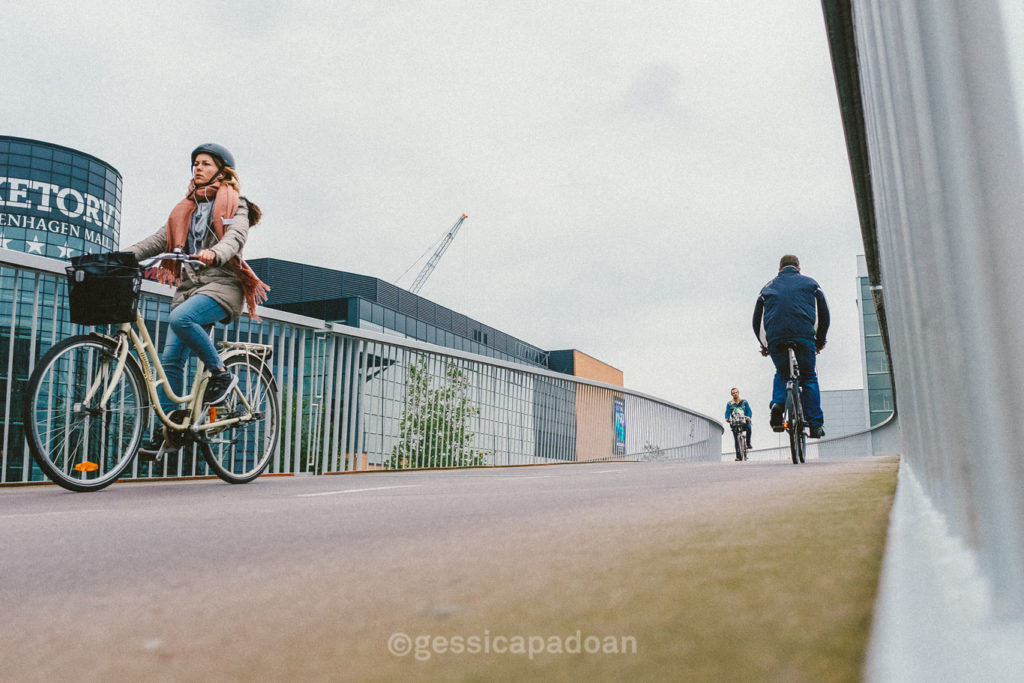 Ciclista su pista ciclabile urbana a Copenaghen, con prospettiva dal basso e passanti in movimento.