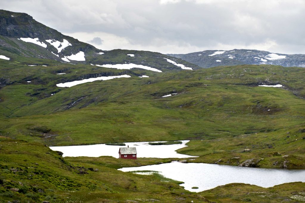 Casetta rossa su prato verde tra montagne innevate e laghi alpini nella regione di Hardangervidda, Norvegia.