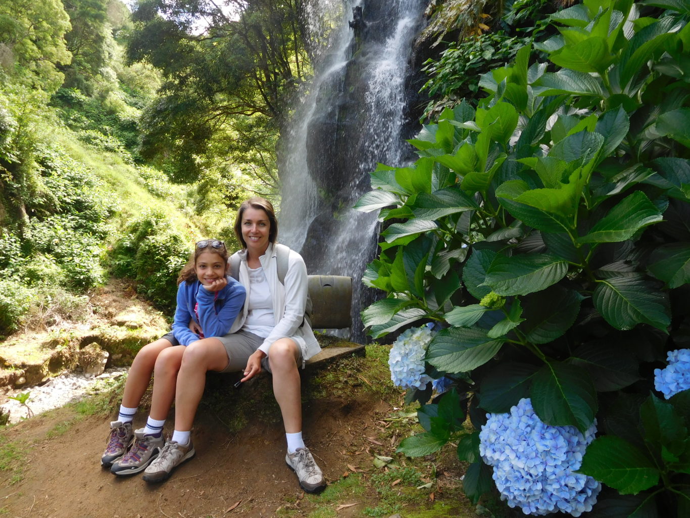 Mamma e figlia in abiti sportivi sedute davanti ad una cascata e vicino ad una pianta di ortensie in fiore