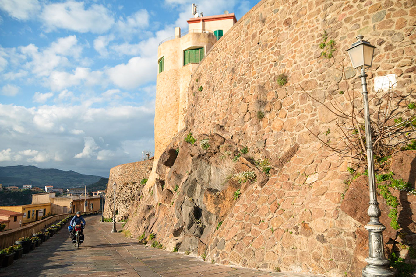 Veduta delle antiche mura di Castelsardo e ciclista che pedala in leggera salita