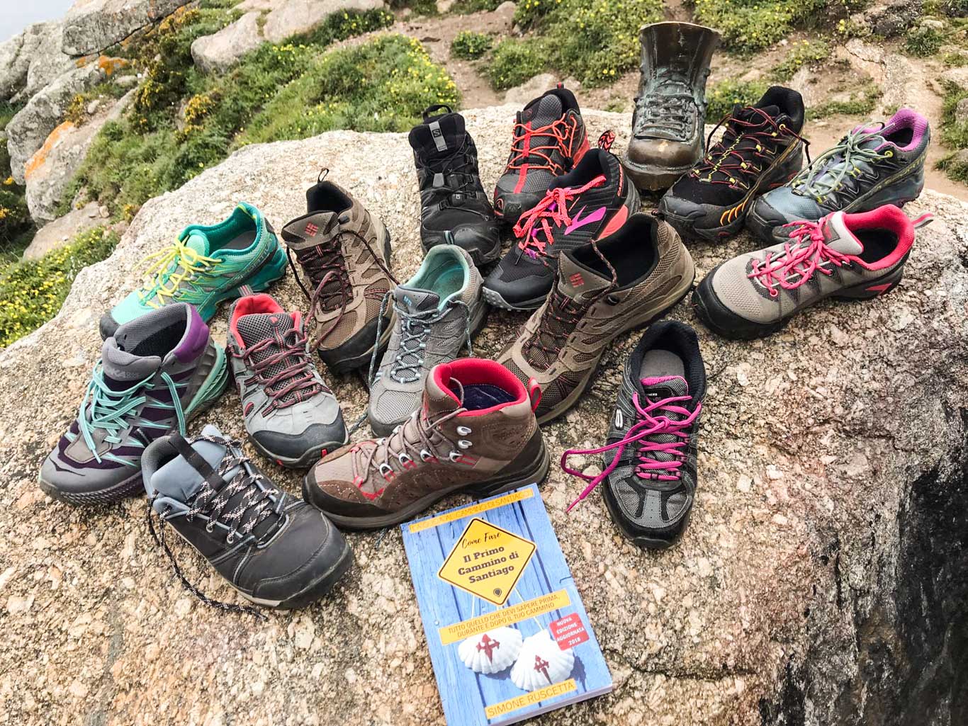 Tantissime scarpe da trekking su una roccia fotografate insieme a un libro sul cammino di Santiago.