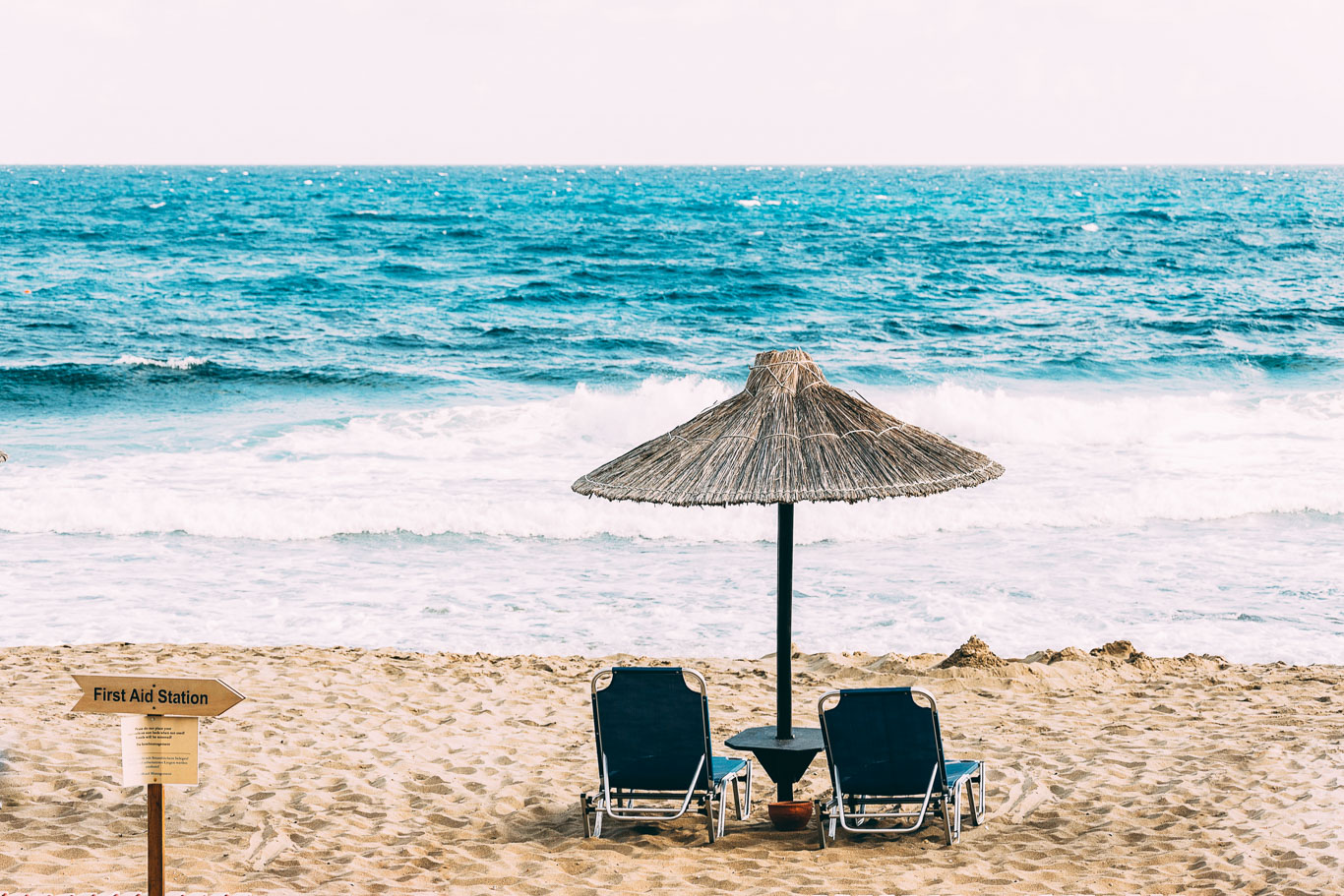 Spiaggia con mare con onde, un ombrellone di paglia e due sdraio blu