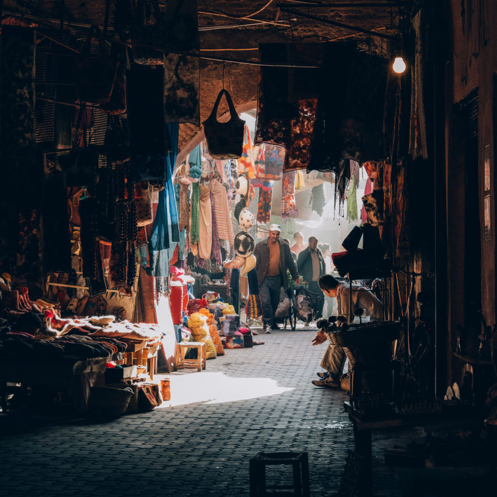 Corridoio interno del suq di Marrakech, tappeti colorati e artigianato appesi, luce filtrata e commercianti marocchini