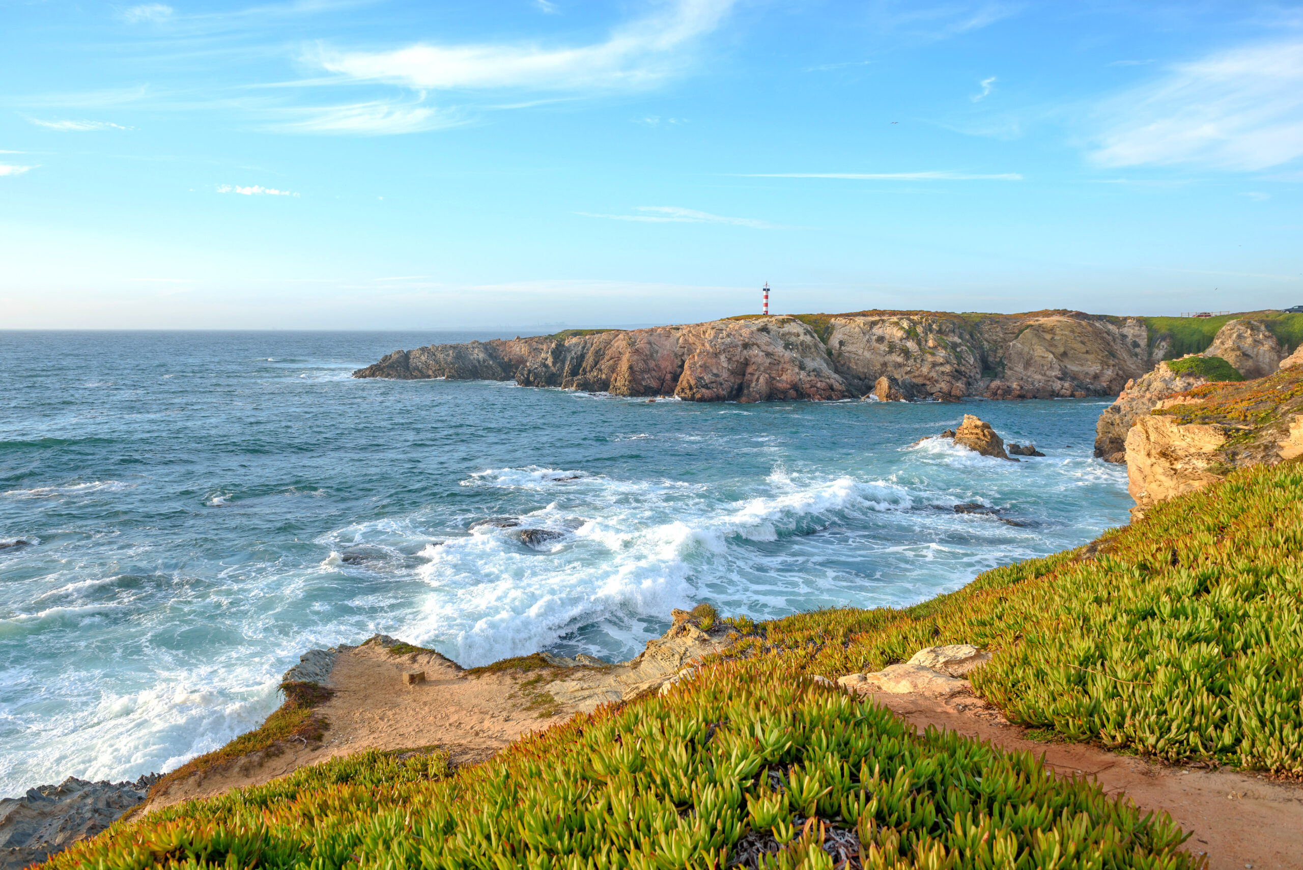 Sentiero della Rota Vicentina con vista su oceano e scogliere con un faro in lontananza