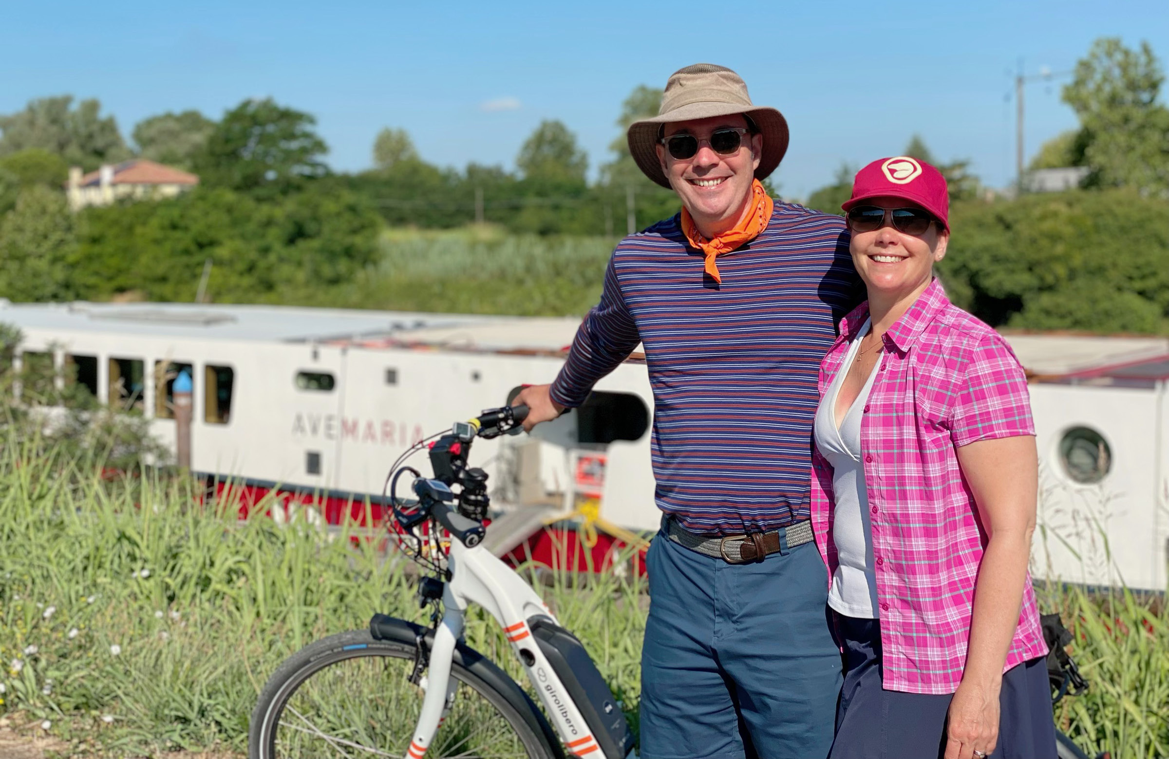 Man and woman embracing and smiling with electric bike and behind Avemaria boat on river.