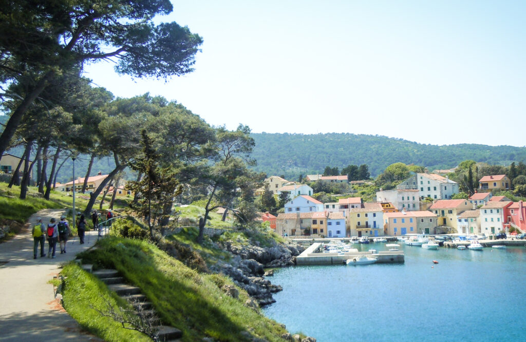 Isola di Lussino in Croazia con gruppo di camminatori a sinistra e mare con case colorate che si affacciano sulla destra.