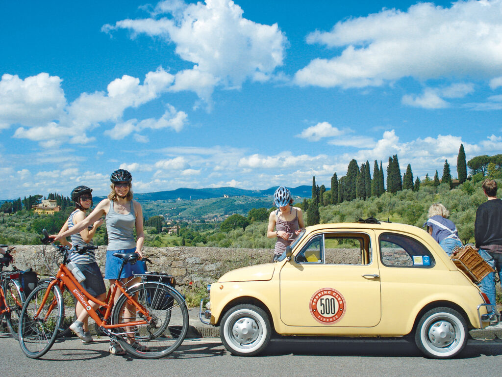 Cyclists posing next to a vintage Fiat 500 with mountain views, northern Italy