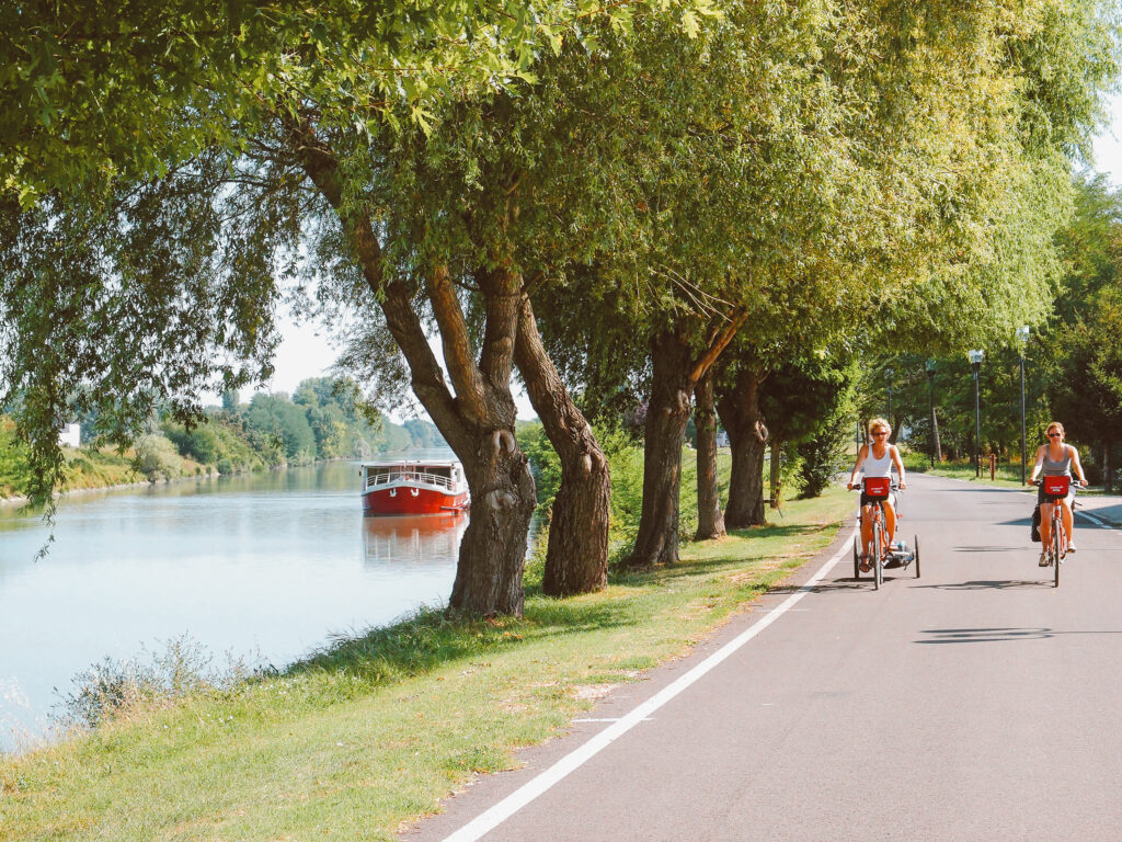 Cyclist riding along a tree-lined canal path with aAve Maria red barge in the background, Veneto waterways